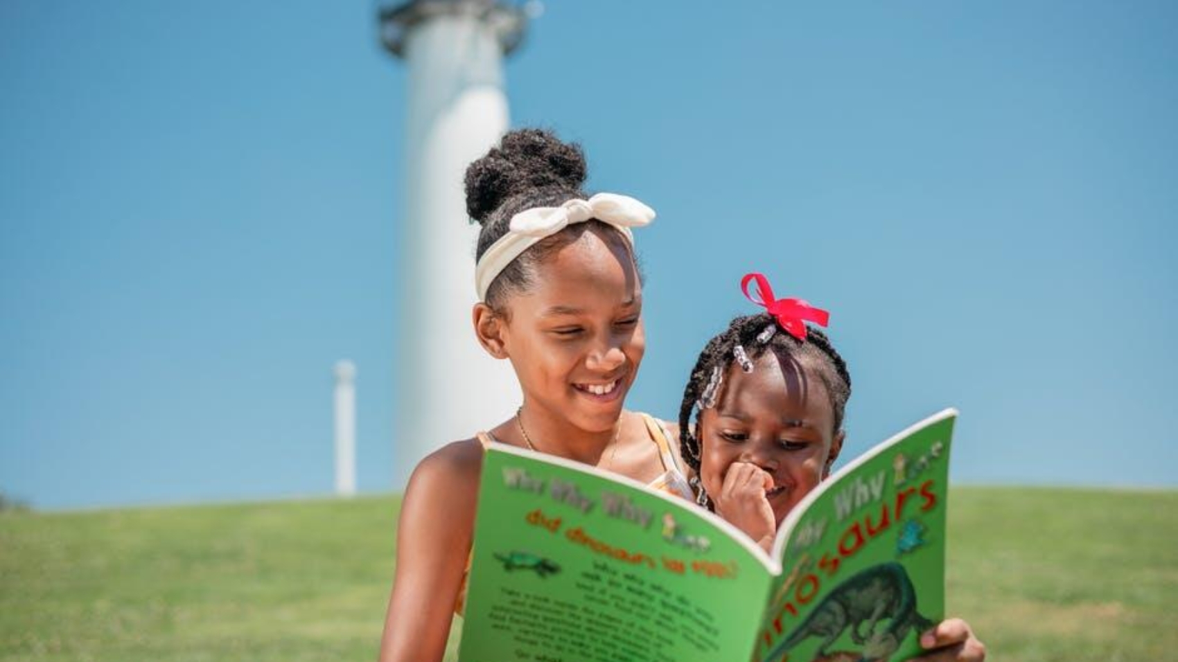 Two young girls read a bright green children's book together on a sunny hill, with a lighthouse in the background.